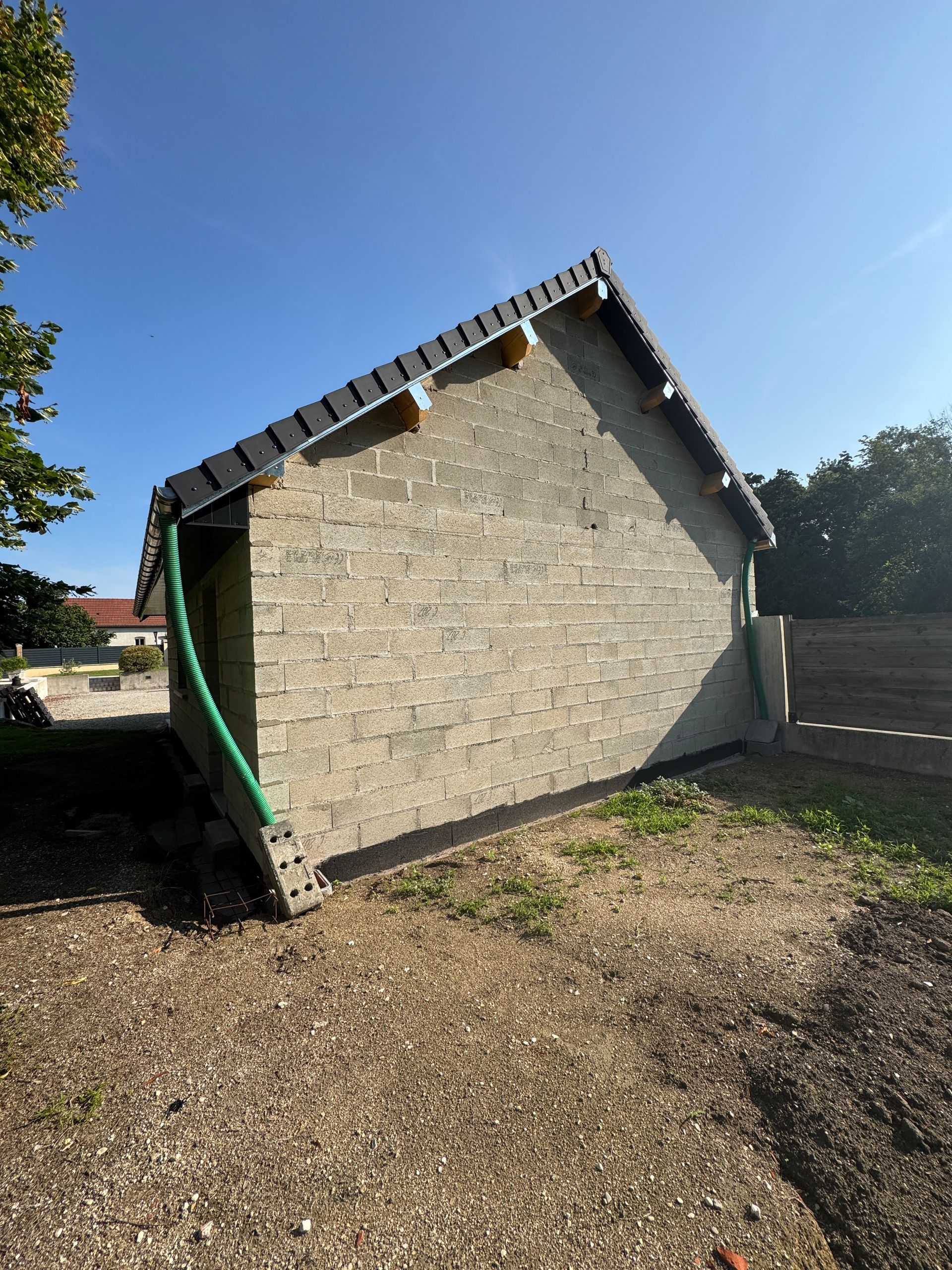 Bâtiment en pierre grise avec un toit de tuiles noires, sur un terrain en gravier avec un tuyau vert et un ciel bleu.