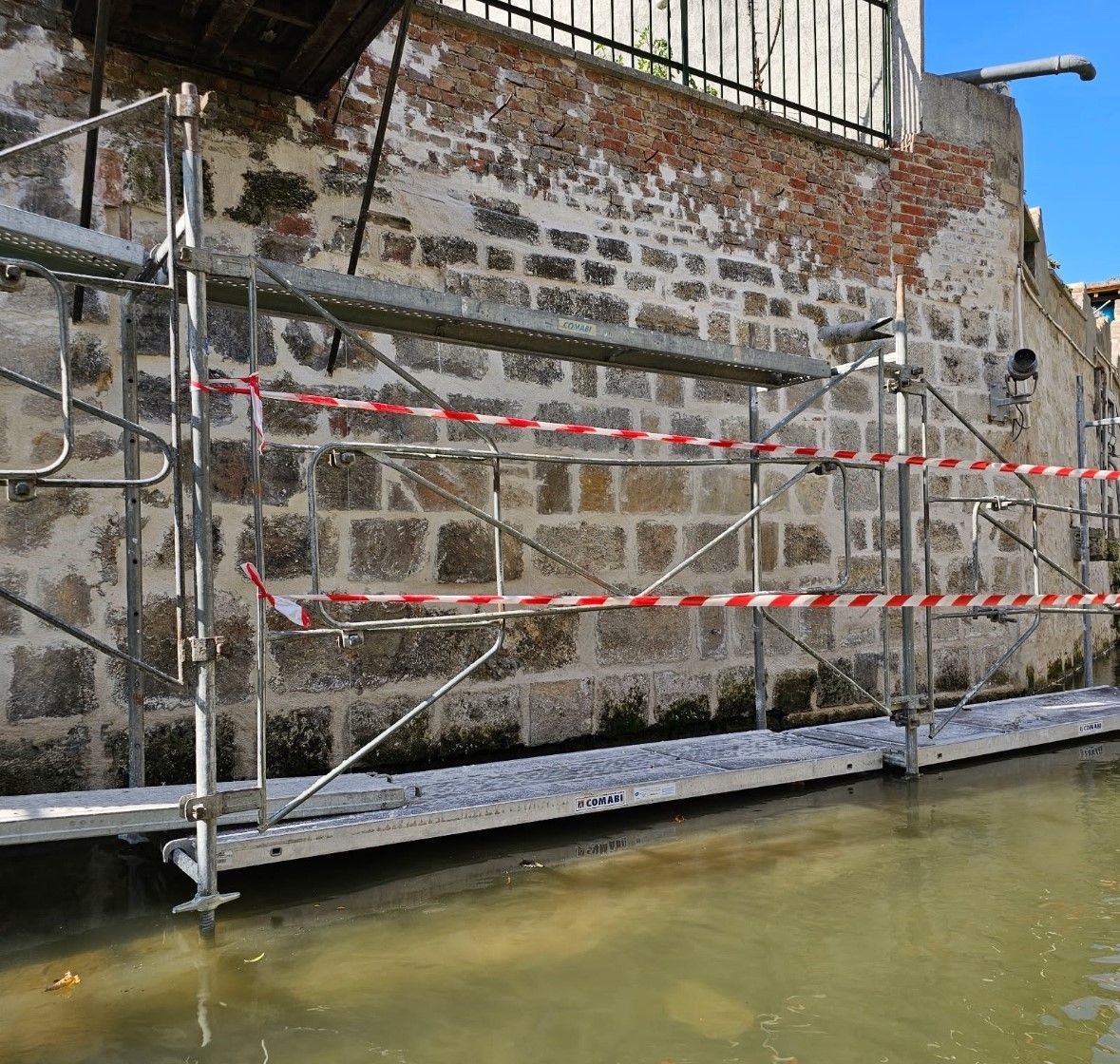 Échafaudage sur un mur de briques au-dessus d'un canal, avec ruban d'avertissement rouge et blanc.