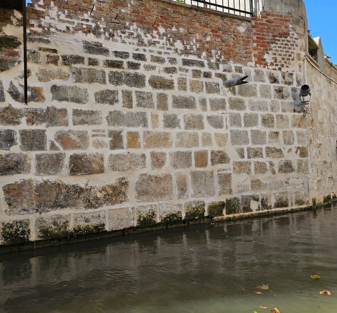 Mur de pierre au bord de l'eau. Quelques feuilles flottent sur l'eau. Au sommet, on aperçoit une clôture en briques et en fer.