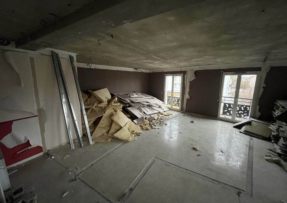 Chambre en travaux avec débris et matériaux apparents. Deux fenêtres, sol gris, isolation beige.