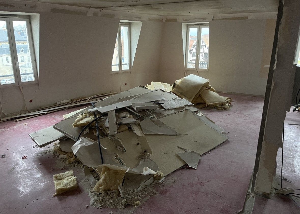 Chambre en mauvais état, avec des débris amoncelés au sol. Moquette rose, fenêtres et murs partiellement démolis sont visibles.