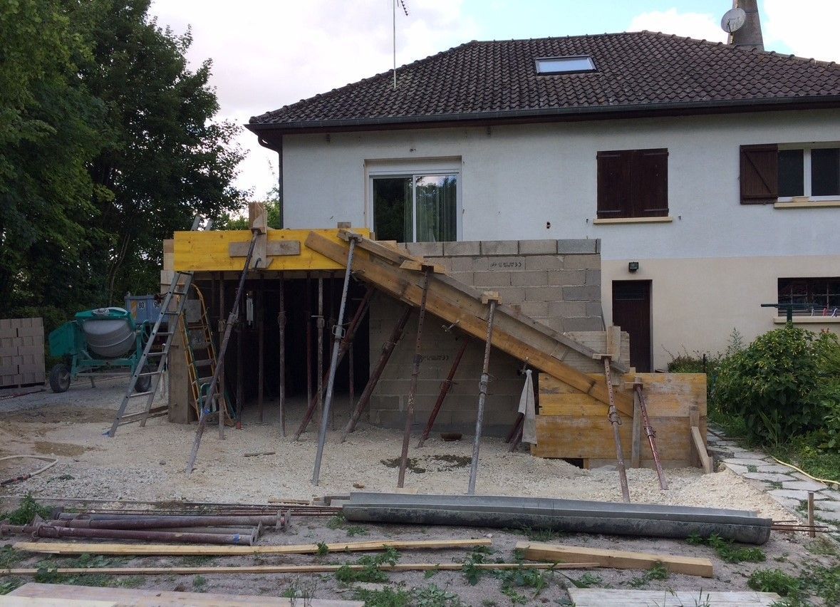 Chantier de construction : une maison avec des fondations en béton, des supports en bois et une rampe menant vers le haut.