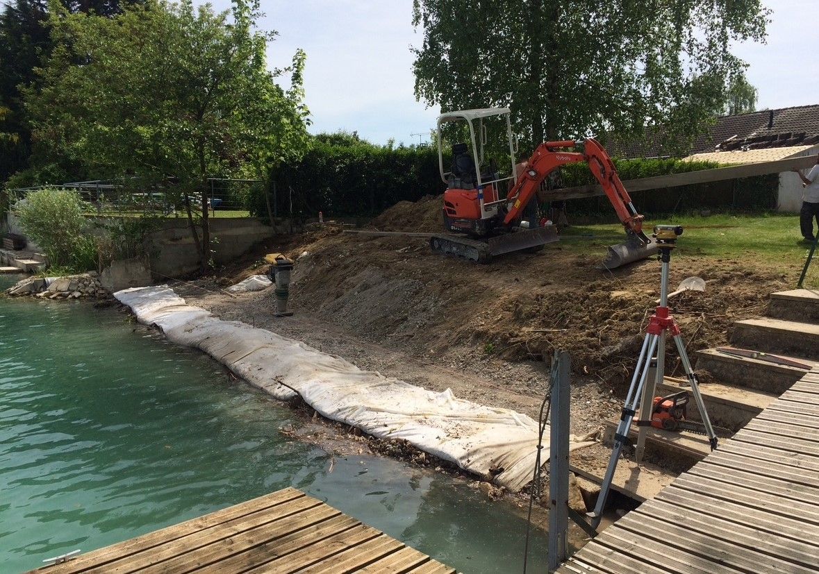 Une excavatrice orange travaillant sur un rivage, à côté d'un quai et d'une eau turquoise.