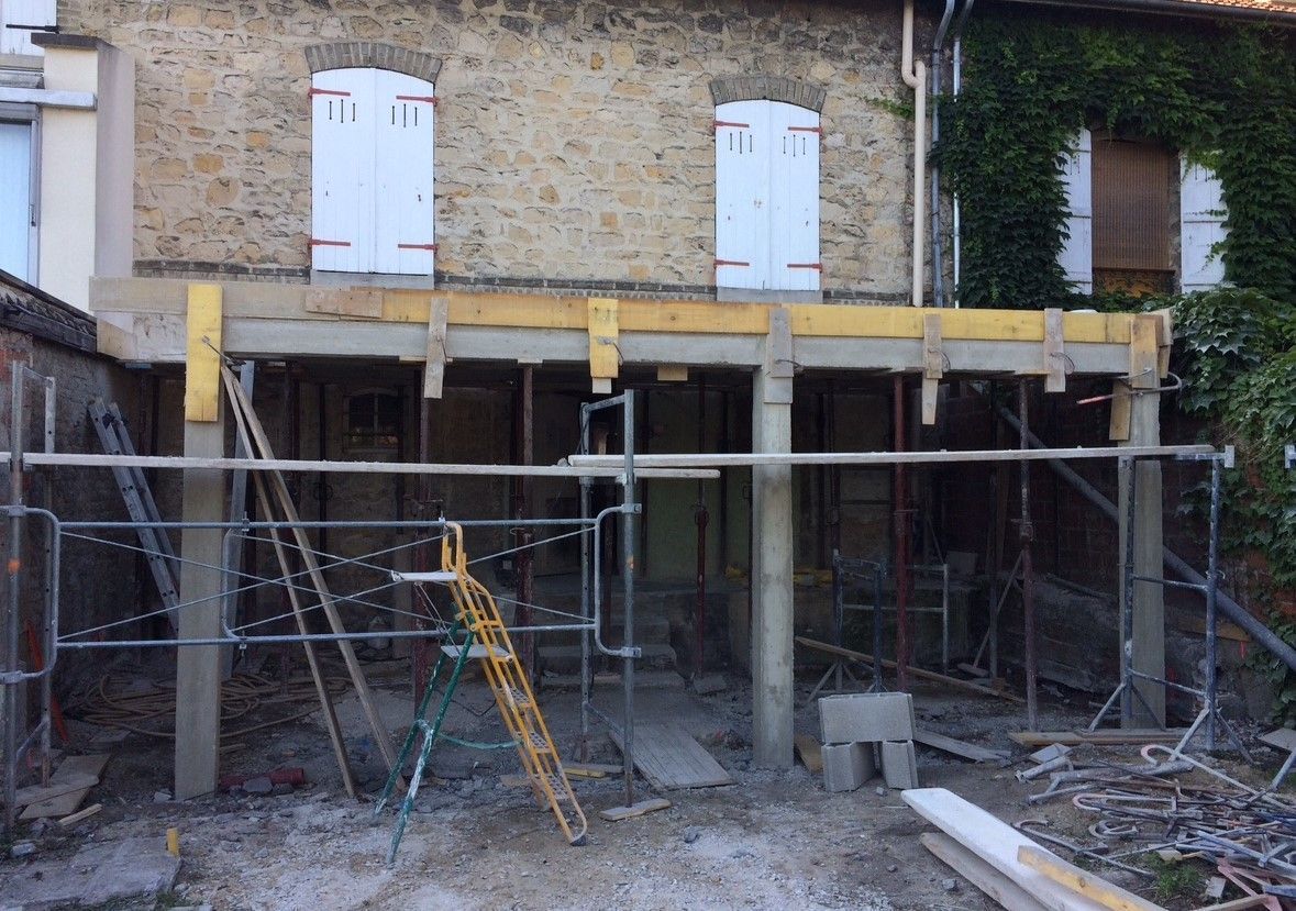 Chantier : piliers en béton soutenant un nouveau porche, avec un bâtiment en pierre en arrière-plan.