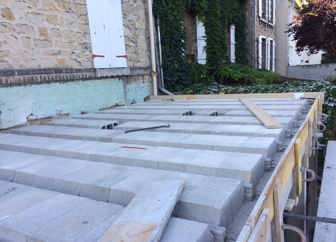 Chantier : pose de blocs de béton pour une structure, supports et ossature en bois, à proximité d'un bâtiment en pierre.