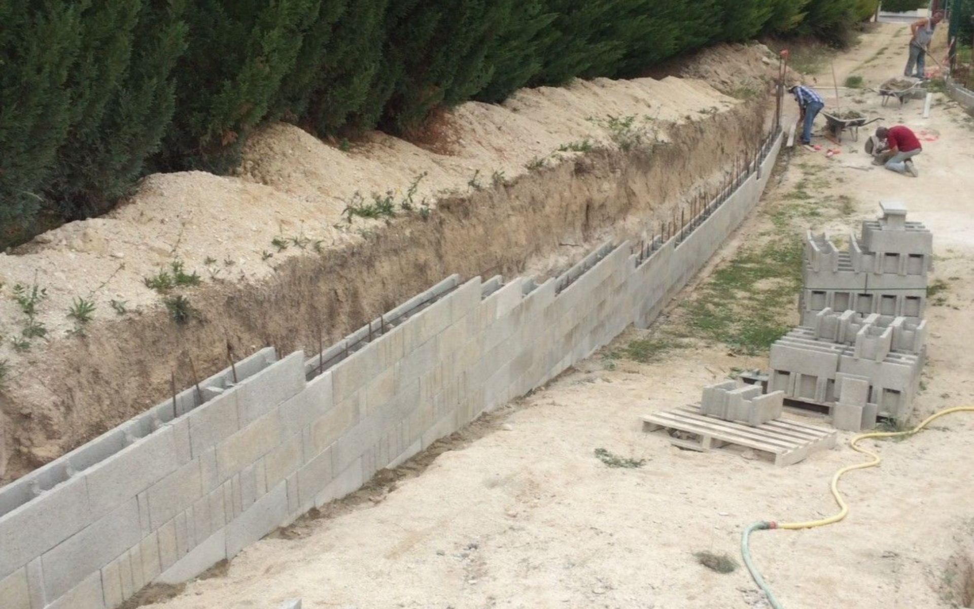 Chantier de construction : des ouvriers construisent un mur de soutènement en blocs de béton le long d'une tranchée, avec une rangée d'arbres en arrière-plan.