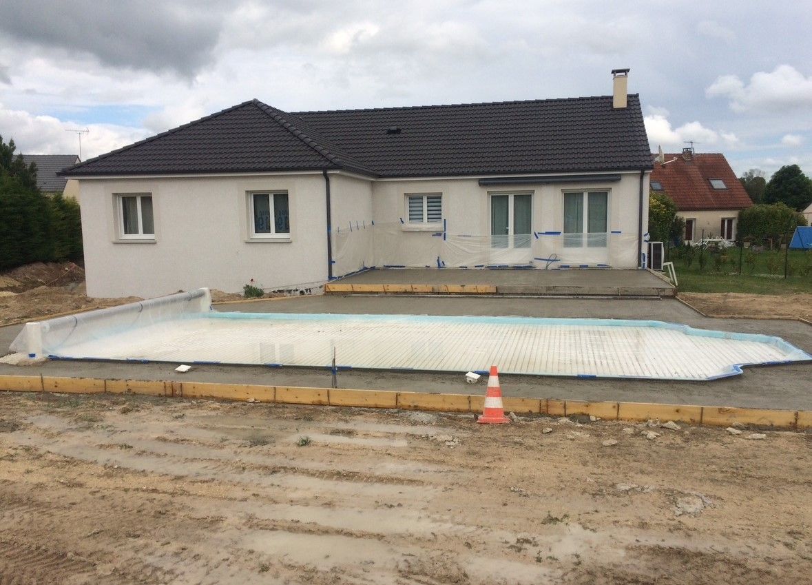 Maison avec une base de piscine nouvellement construite ; une maison aux murs blancs avec un toit sombre dans une cour en terre.