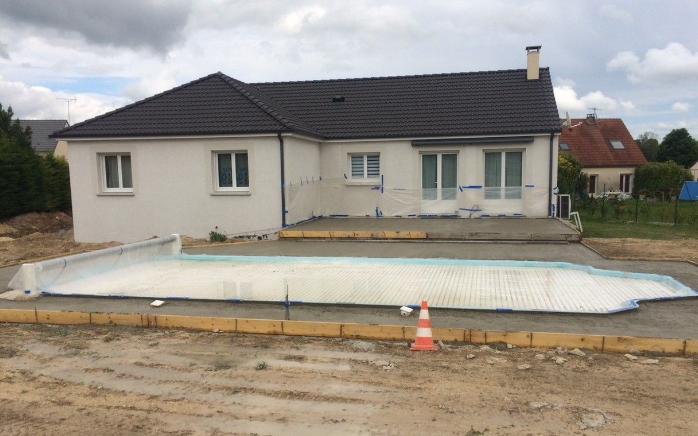 Maison en construction avec patio en béton, fondations de piscine et cône de signalisation orange.