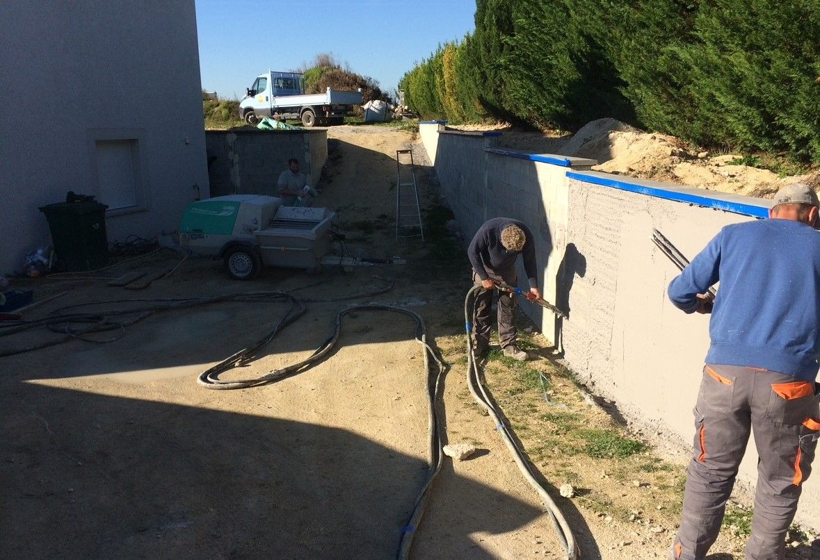 Des ouvriers du bâtiment plâtraient un mur à l'extérieur. Une machine et un camion sont visibles à l'arrière-plan.