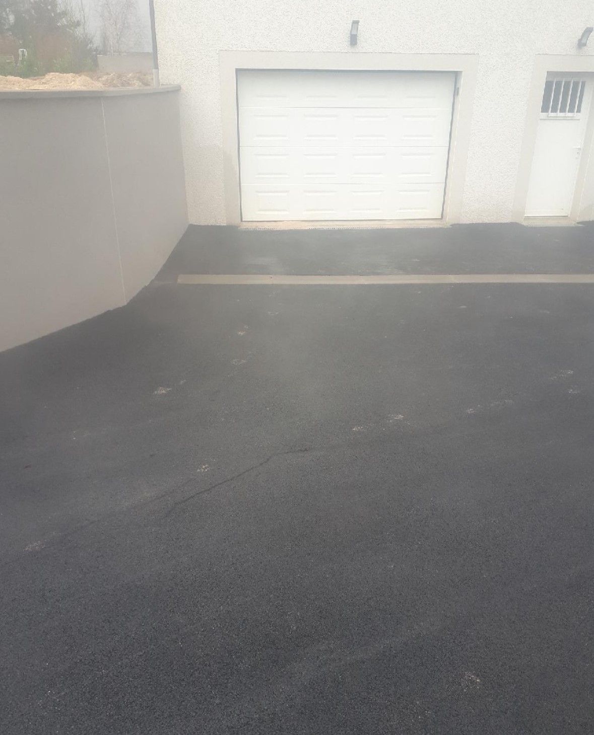 Allée en asphalte noir menant à un garage et une porte blancs, un mur gris clair se trouve sur la gauche.