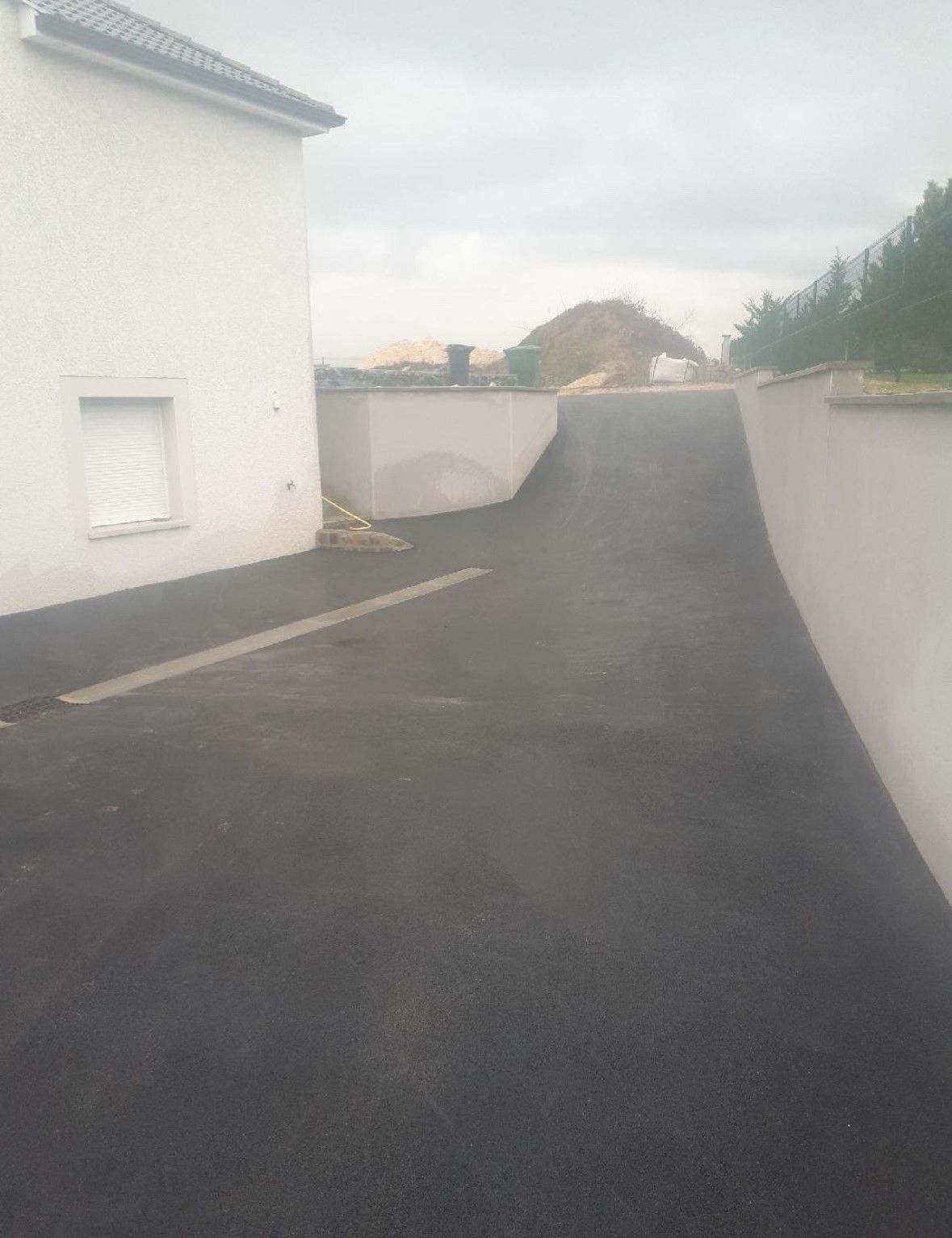 L'allée asphaltée monte en pente entre des murs blancs, à côté d'un bâtiment blanc sous un ciel couvert.