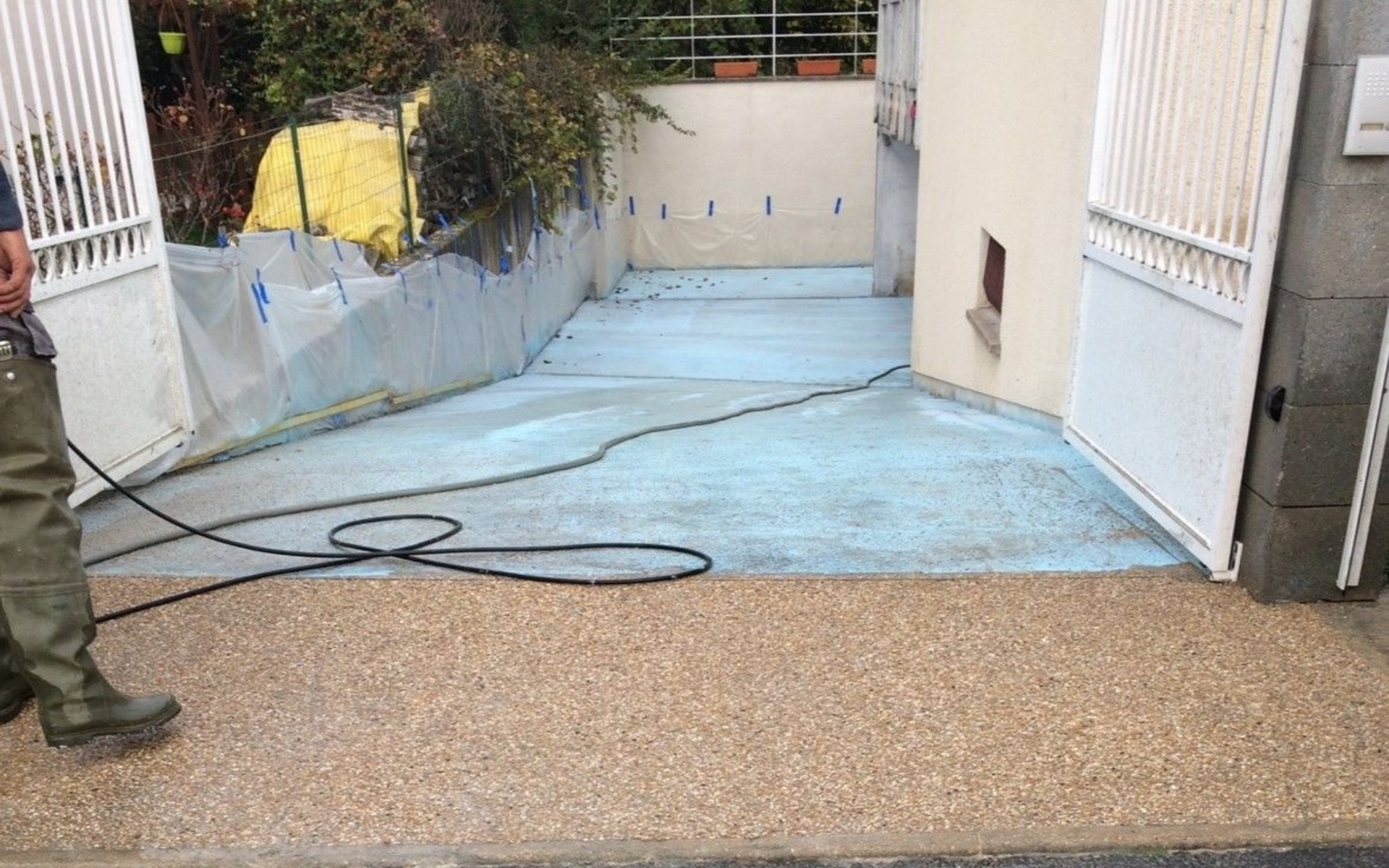 Un homme en bottes de travail se tient près d'une allée en béton bleu clair. Portail blanc ouvert.