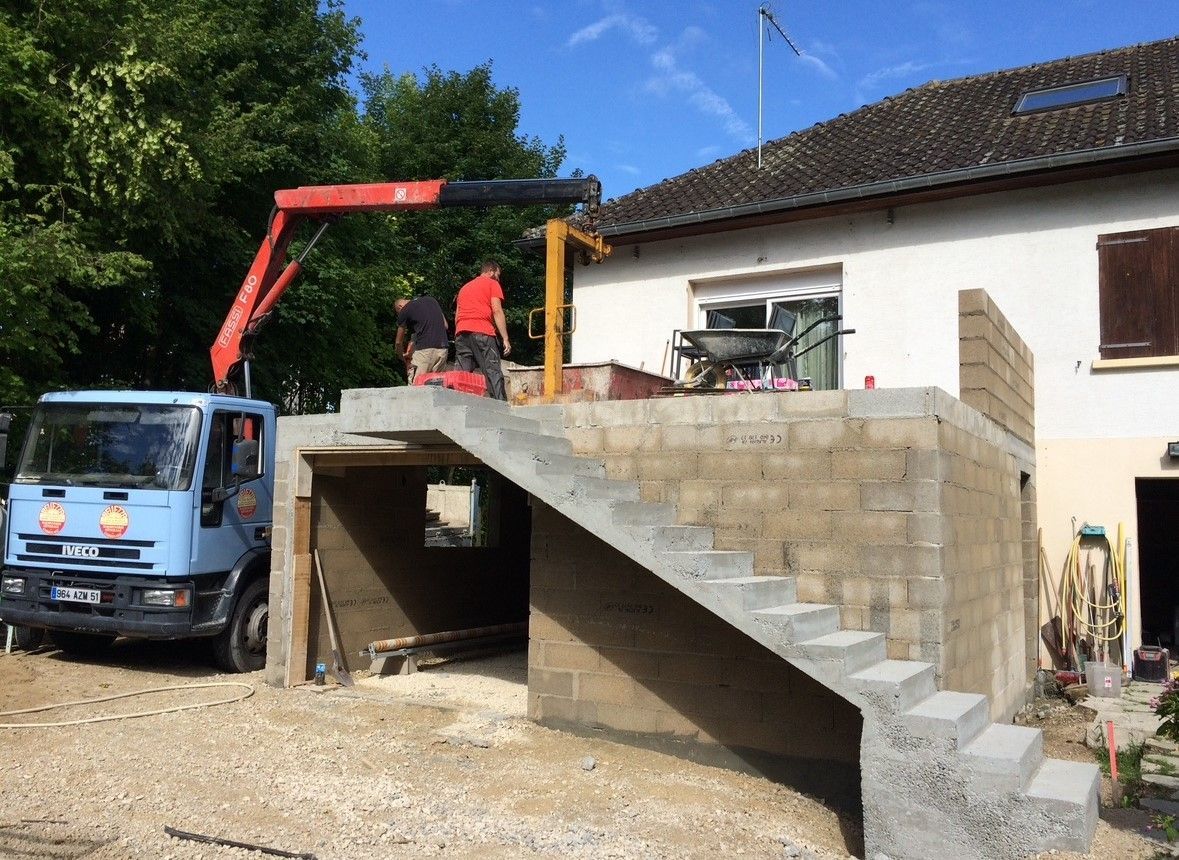 Chantier de construction : camion avec grue soulevant des matériaux sur une structure en parpaings, ouvriers en chemises rouges, escaliers et maison en arrière-plan.