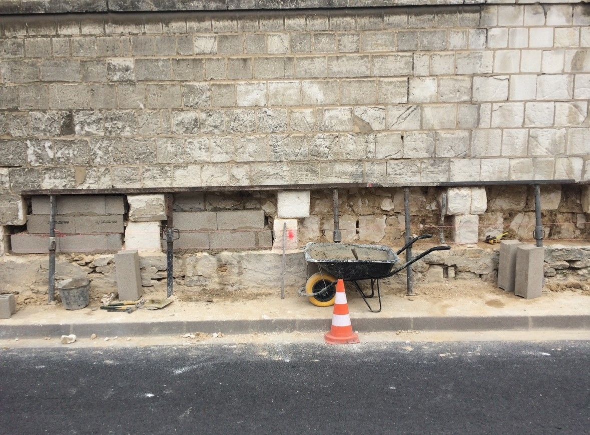 Chantier : mur en réparation, avec une brouette, un cône et des blocs de béton.