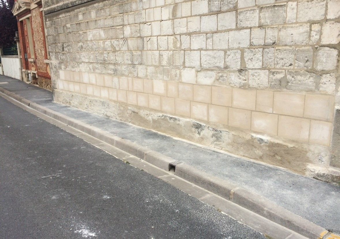 Mur de briques grises avec des carreaux beiges en bas, le long d'un trottoir gris et d'une gouttière dans une rue pavée.
