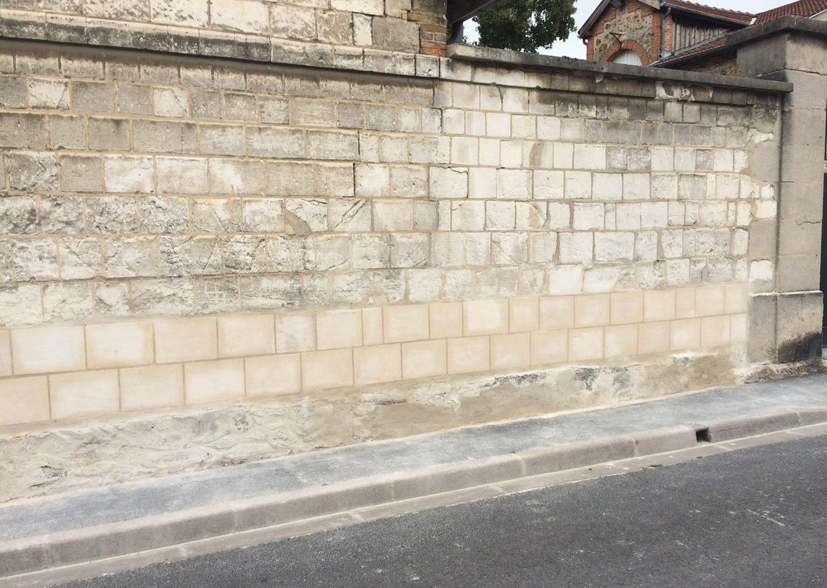 Mur de briques dans une rue, patiné et décoloré, avec des blocs de couleur plus claire près de la base.