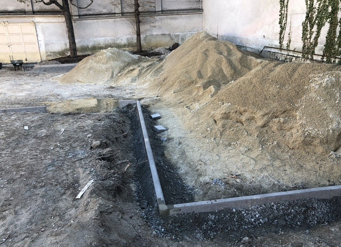 Tas de terre près d'une bordure rectangulaire en béton, potentiellement pour un jardin, avec un bâtiment en arrière-plan.