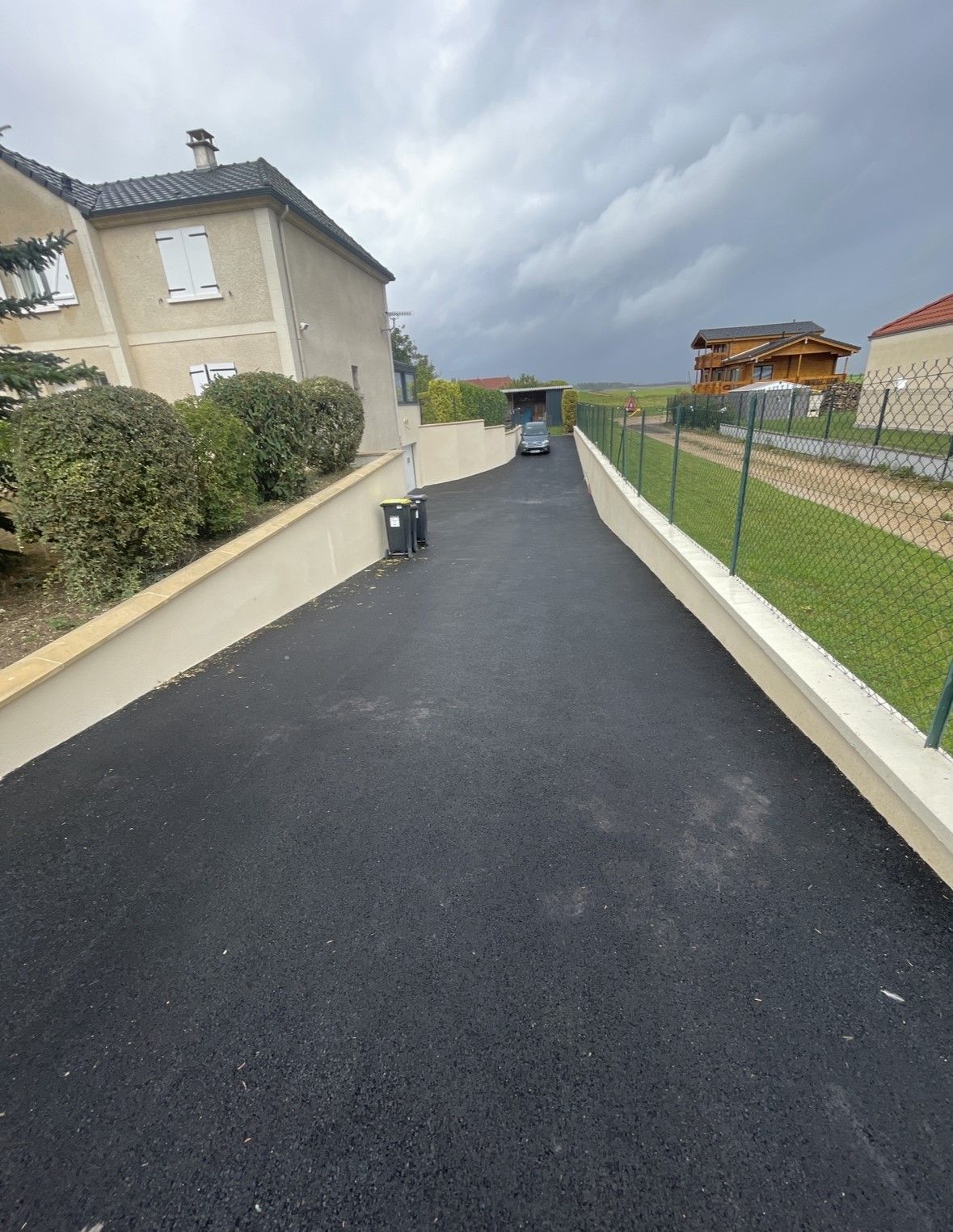 Allée asphaltée entre deux murets, menant à une maison par temps nuageux.