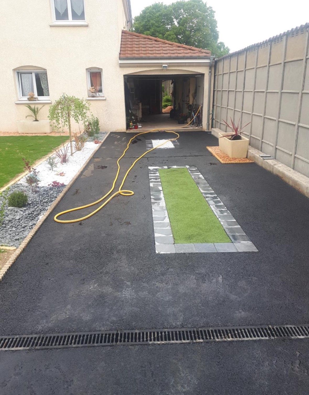 Allée asphaltée avec garage, petite pelouse et parterres de fleurs.