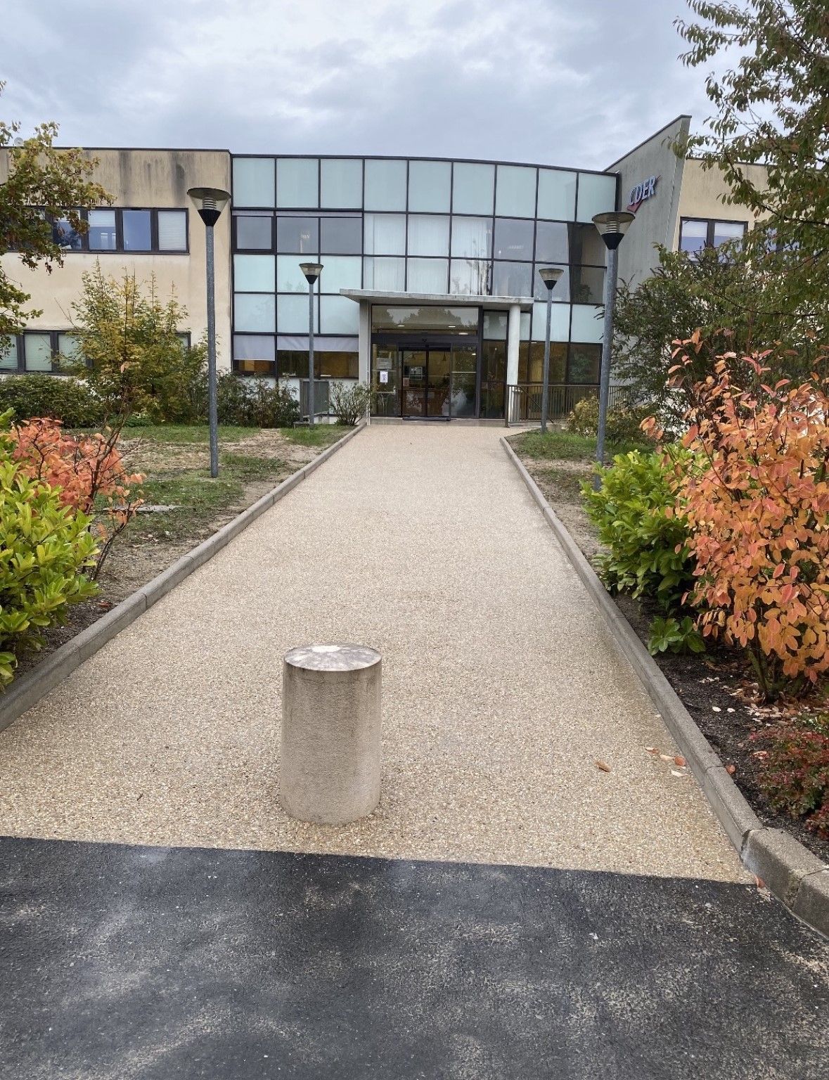 Entrée du bâtiment avec façade vitrée, allée de gravier et feuillage d'automne.