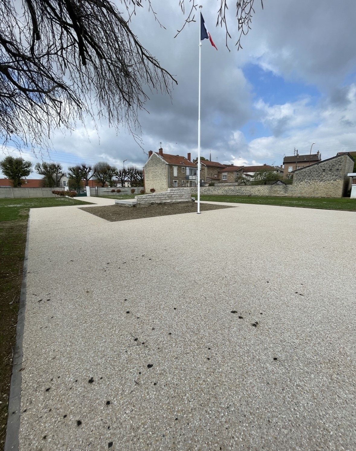 Place en gravier avec le drapeau français sur un mât. Bâtiments en pierre et ciel nuageux en arrière-plan.