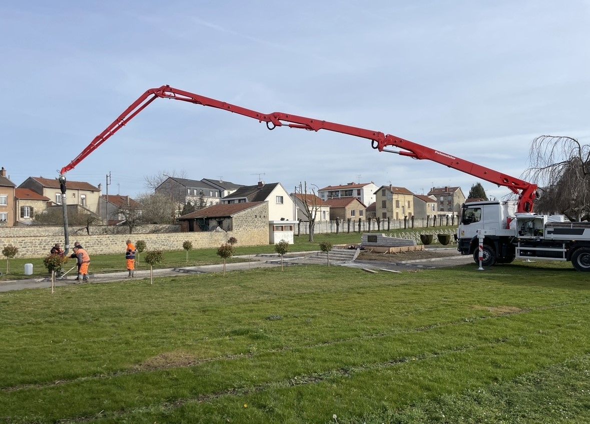 Un camion-pompe à béton rouge s'étendant sur une zone herbeuse, des ouvriers sont présents ; des bâtiments en arrière-plan.