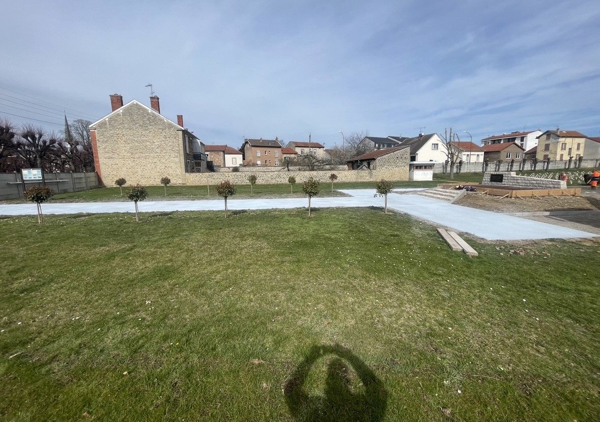 Parc herbeux avec chemin pavé et petits arbres, bâtiments en pierre en arrière-plan sous un ciel bleu.