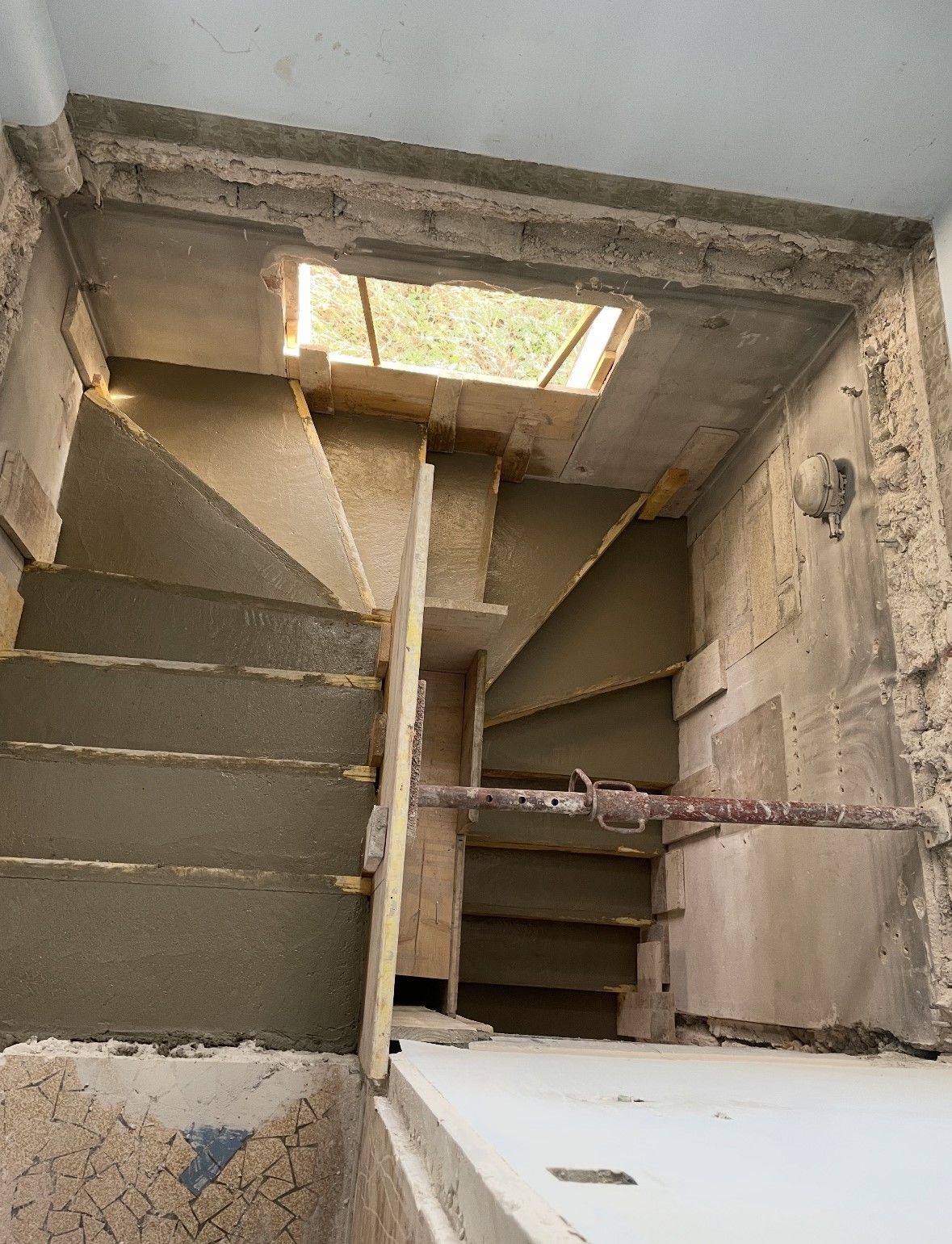 Un escalier étroit et en colimaçon descendant, surmonté d'une lucarne. Les murs sont en béton brut.