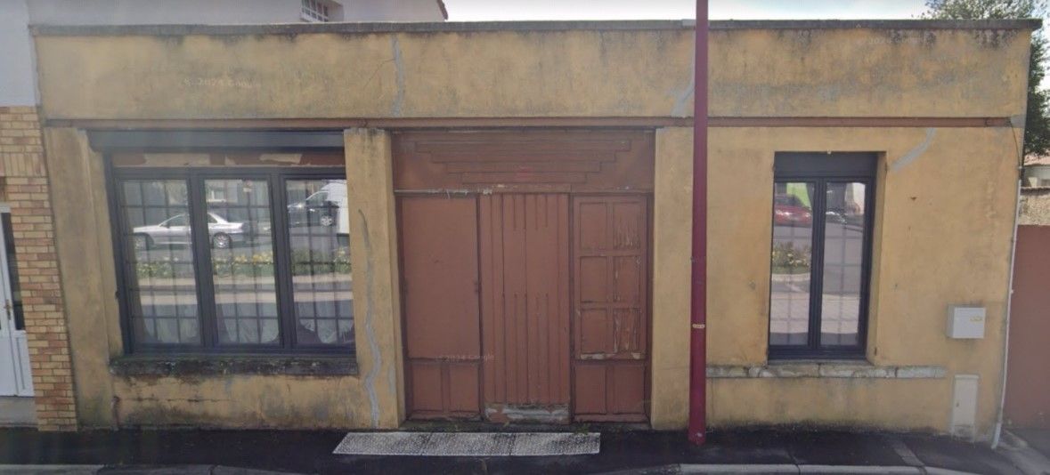 Un bâtiment de couleur beige avec une grande porte marron, deux fenêtres et un poteau rouge devant.