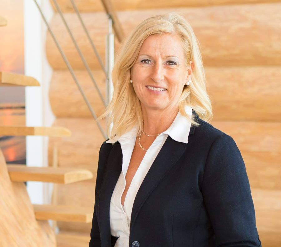 Eine Frau in Anzug und weißem Hemd steht vor einer Holztreppe.