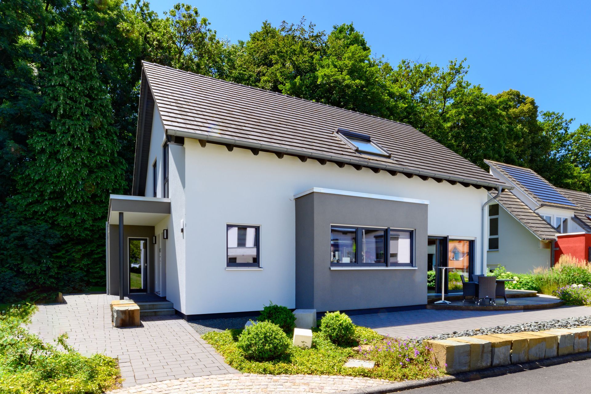 Maison blanche moderne avec un toit ondulé foncé, de grandes fenêtres et un petit jardin paysager sous un ciel bleu.