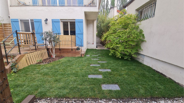 Petit jardin avec pelouse, dalles de pierre, terrasse en bois et maison aux volets bleus.