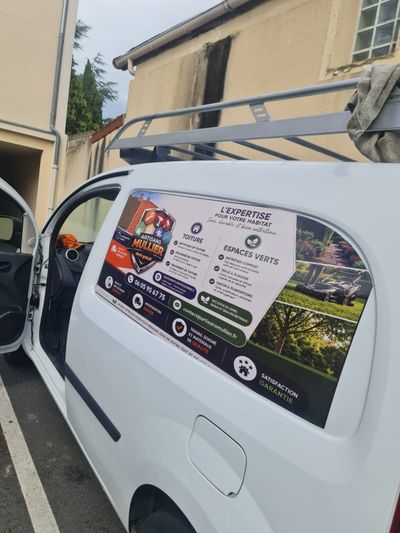 Une camionnette blanche ornée d'autocollants publicitaires est garée à côté d'un bâtiment beige.