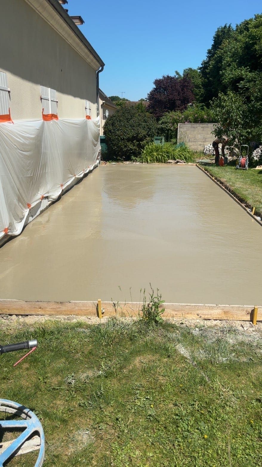 Dalle de béton fraîchement coulée à côté d'une maison, bordée de pelouse et d'un jardin ensoleillé.