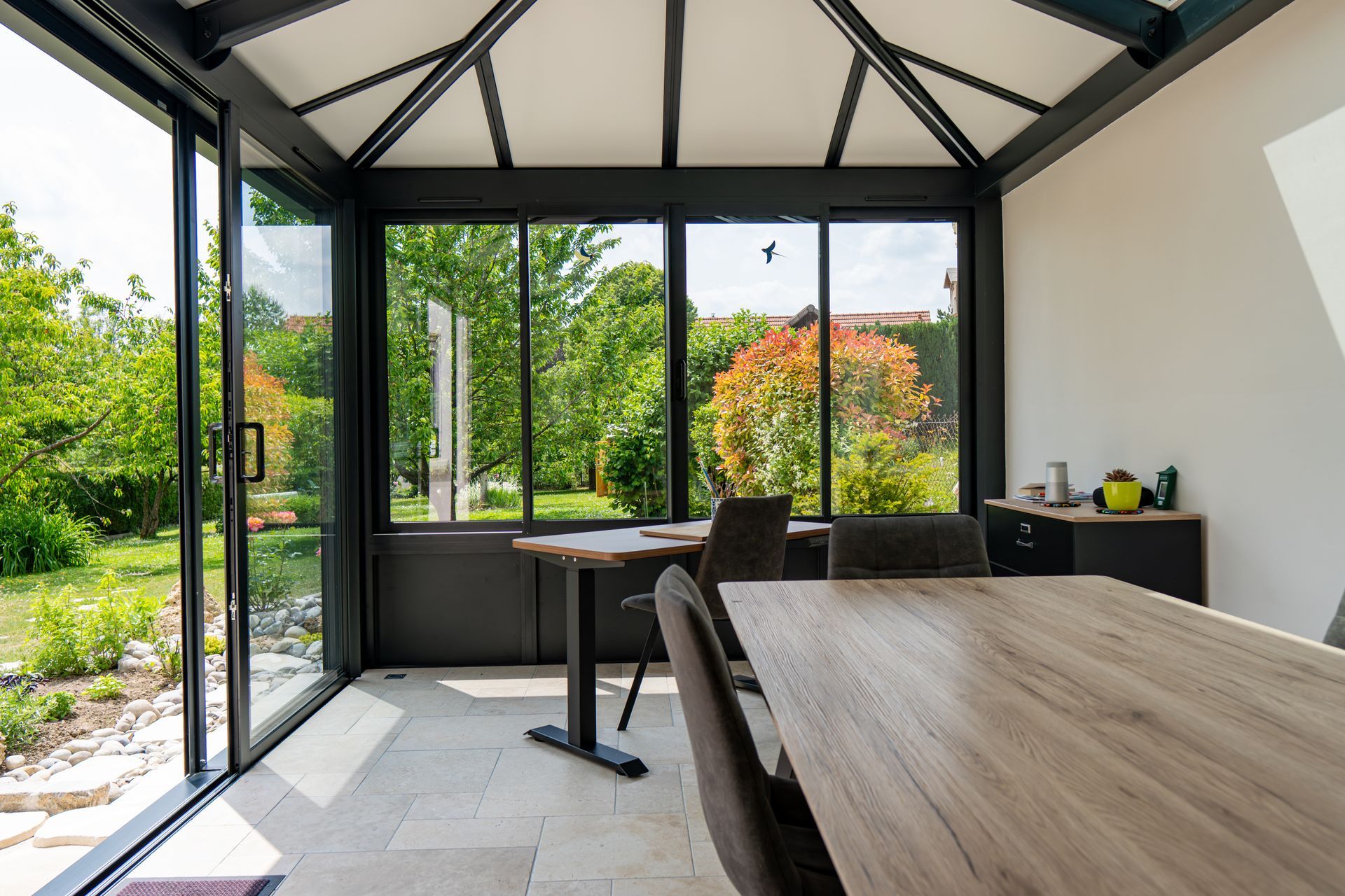 Salle à manger moderne avec de grandes fenêtres, table en bois, chaises noires et vue sur le jardin.
