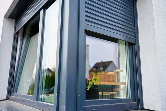 Fenêtre grise moderne avec volets foncés reflétant une cour arrière et une maison voisine.
