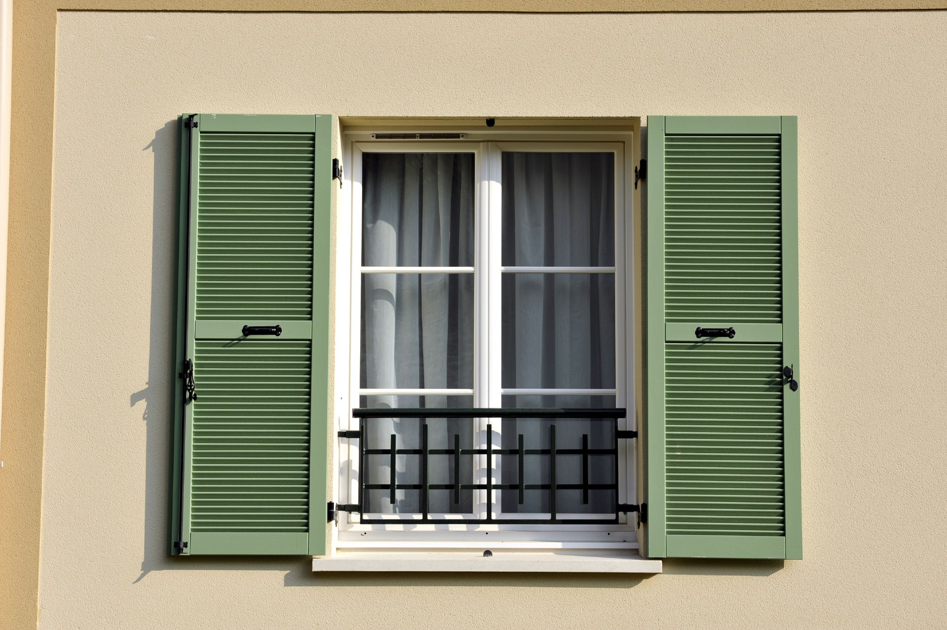 Des volets verts encadrent une fenêtre blanche avec des rideaux et une rambarde en fer forgé noir.