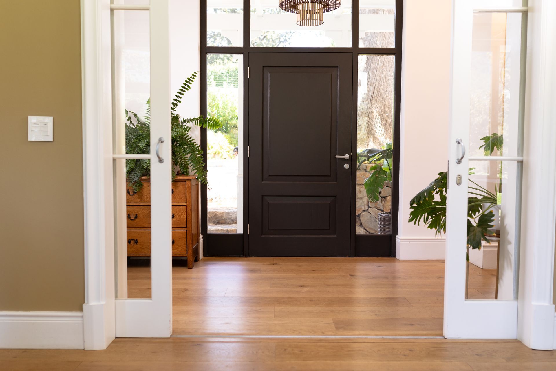 Entrée principale avec une porte à panneaux sombres, un parquet et des plantes en pot visibles à travers les portes-fenêtres ouvertes.