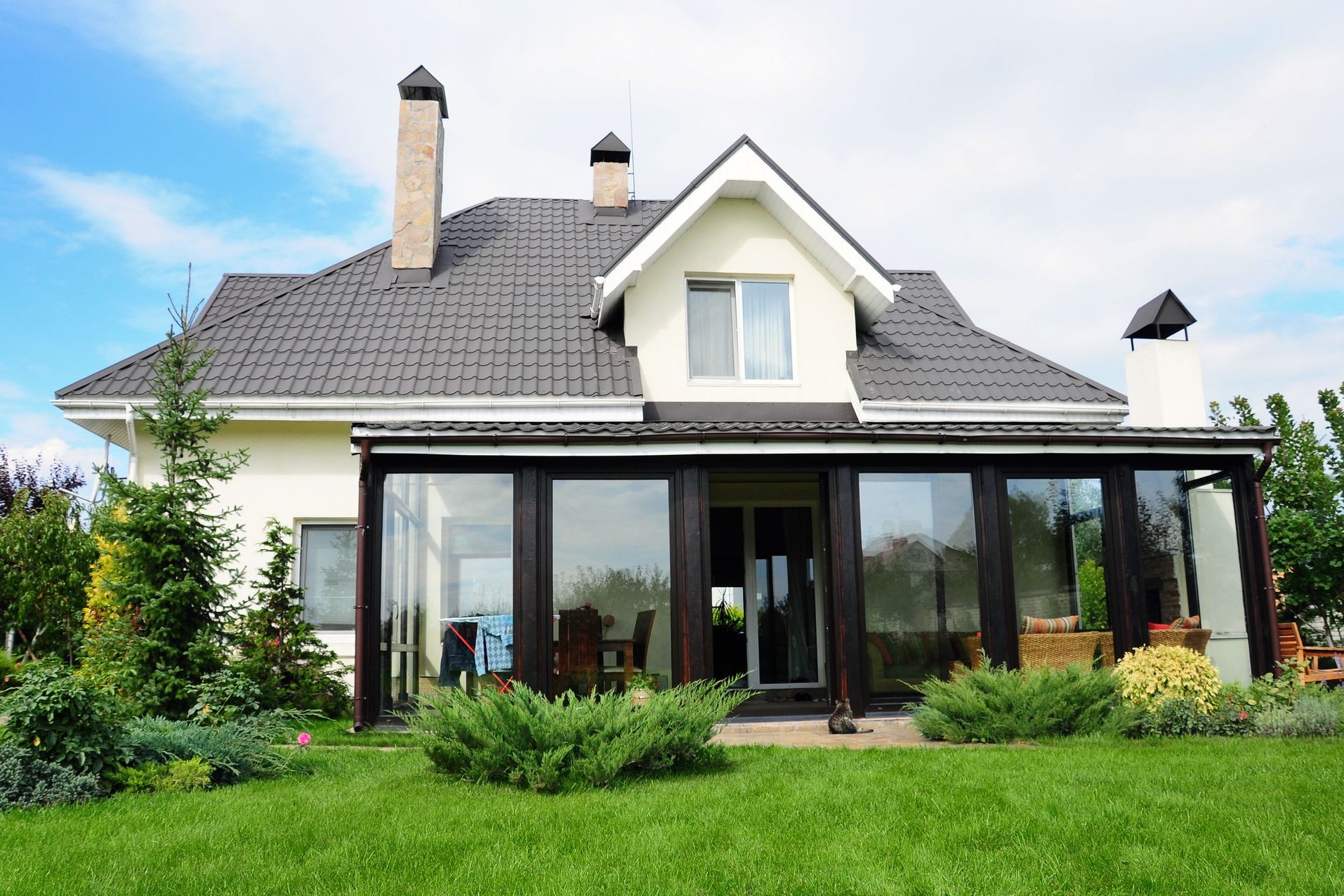 Maison moderne aux murs blancs, au toit gris et dotée d'une véranda vitrée, entourée d'une pelouse et d'un jardin verdoyants.