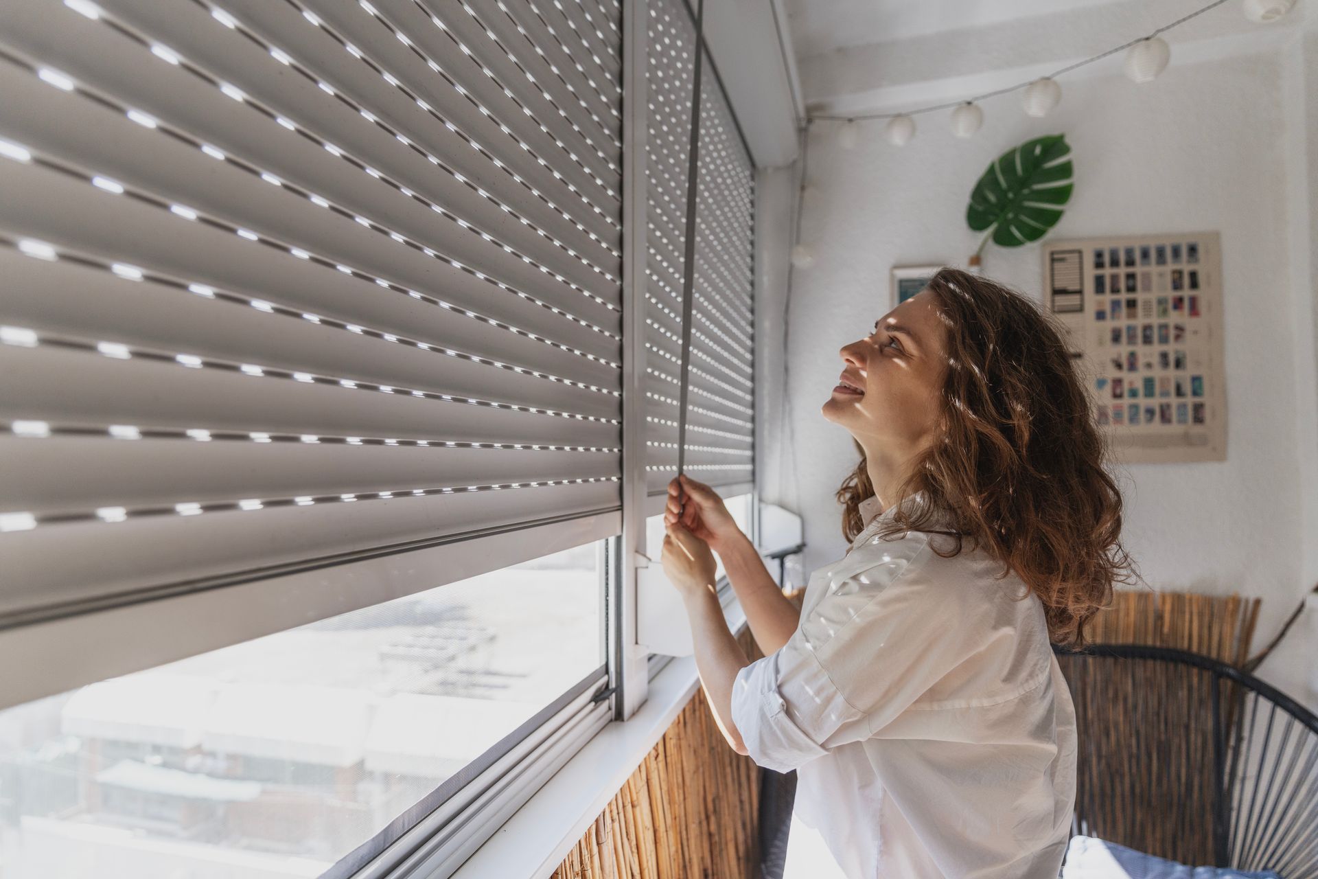Une femme ouvre les stores d'une fenêtre dans une pièce lumineuse aux murs blancs et à la décoration murale en forme de feuille verte.