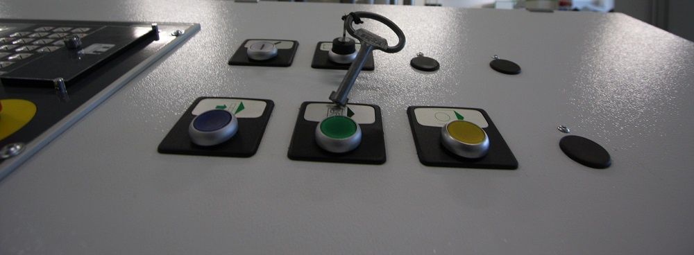 Gros plan sur trois boutons colorés et un levier incurvé sur un panneau de commande gris, avec le bord d'un ordinateur portable visible.