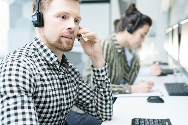 Un agent de centre d'appels, portant un casque, parle à un bureau, un autre employé se trouvant en arrière-plan.
