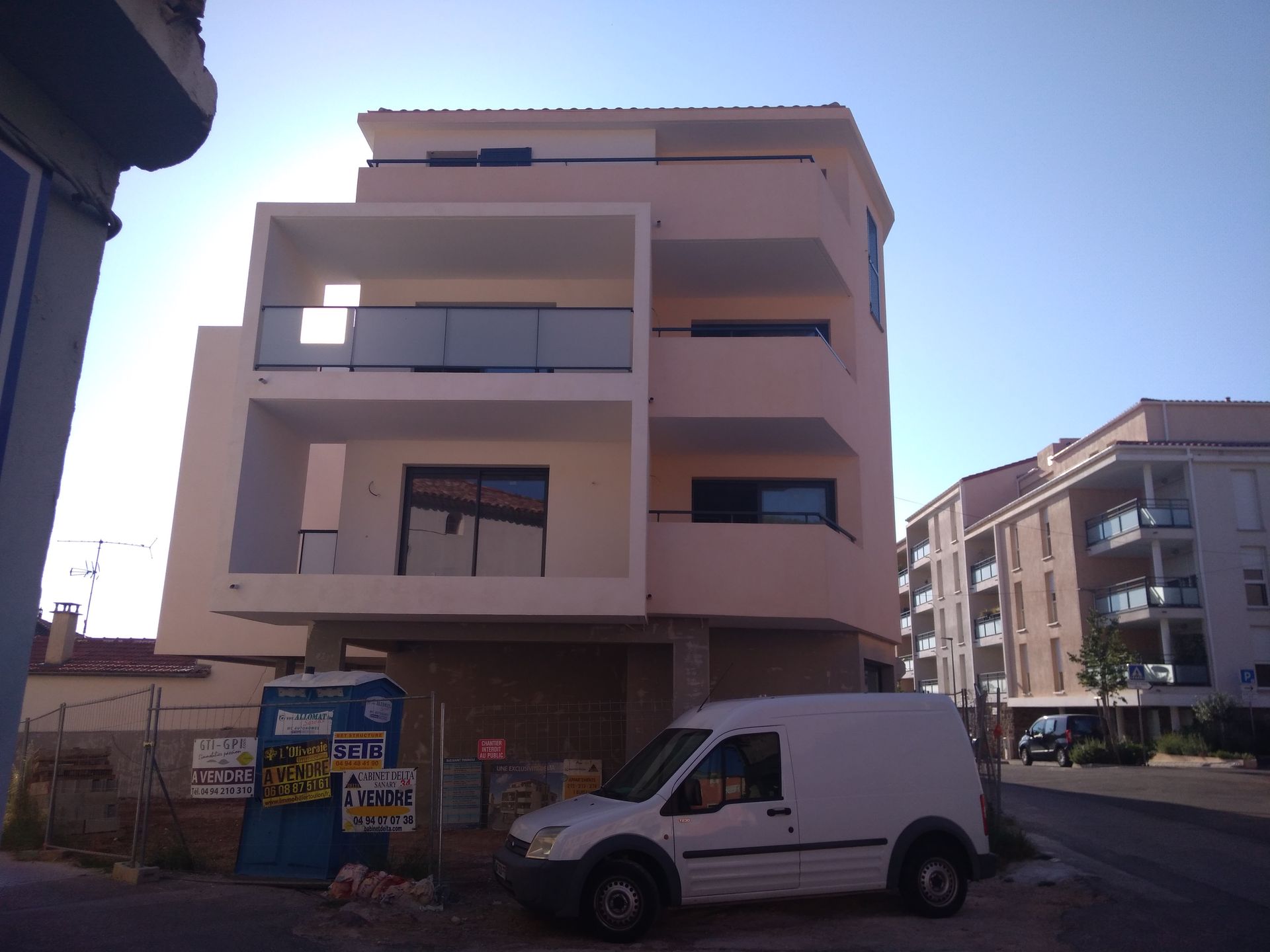 Un immeuble beige à plusieurs étages avec des balcons et une camionnette blanche garée devant par une journée ensoleillée.