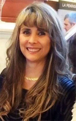 Una mujer sonriente con cabello castaño largo, vestida con una blusa negra texturizada y un collar de cadena dorada.