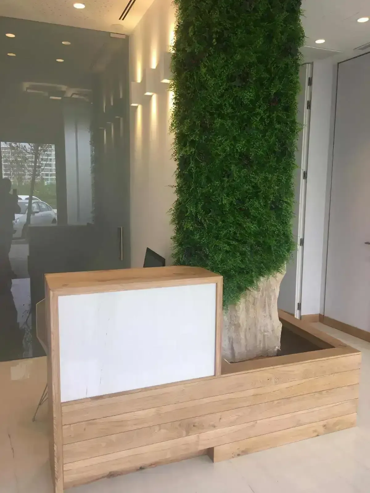 Un mostrador de recepción moderno, fabricado en madera clara con un panel blanco, que incorpora una jardinera
