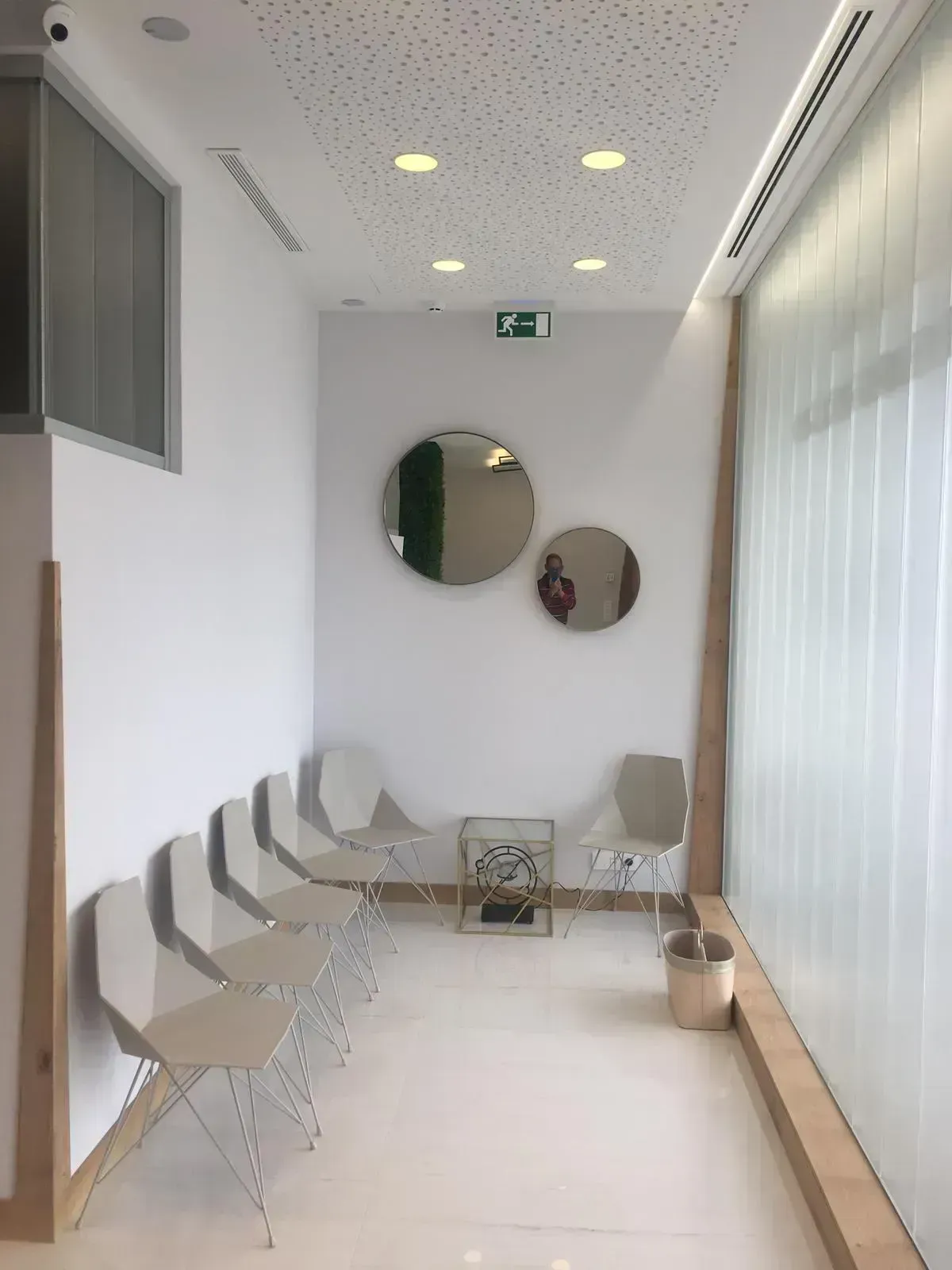 Una sala de espera minimalista con una fila de cinco sillas blancas, dos espejos redondos en una pared blanca