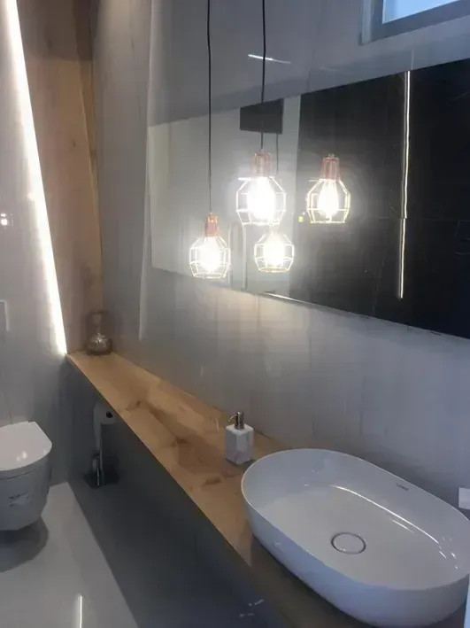 Baño moderno con encimera de madera, lavabo ovalado blanco y tres lámparas colgantes geométricas sobre un espejo.