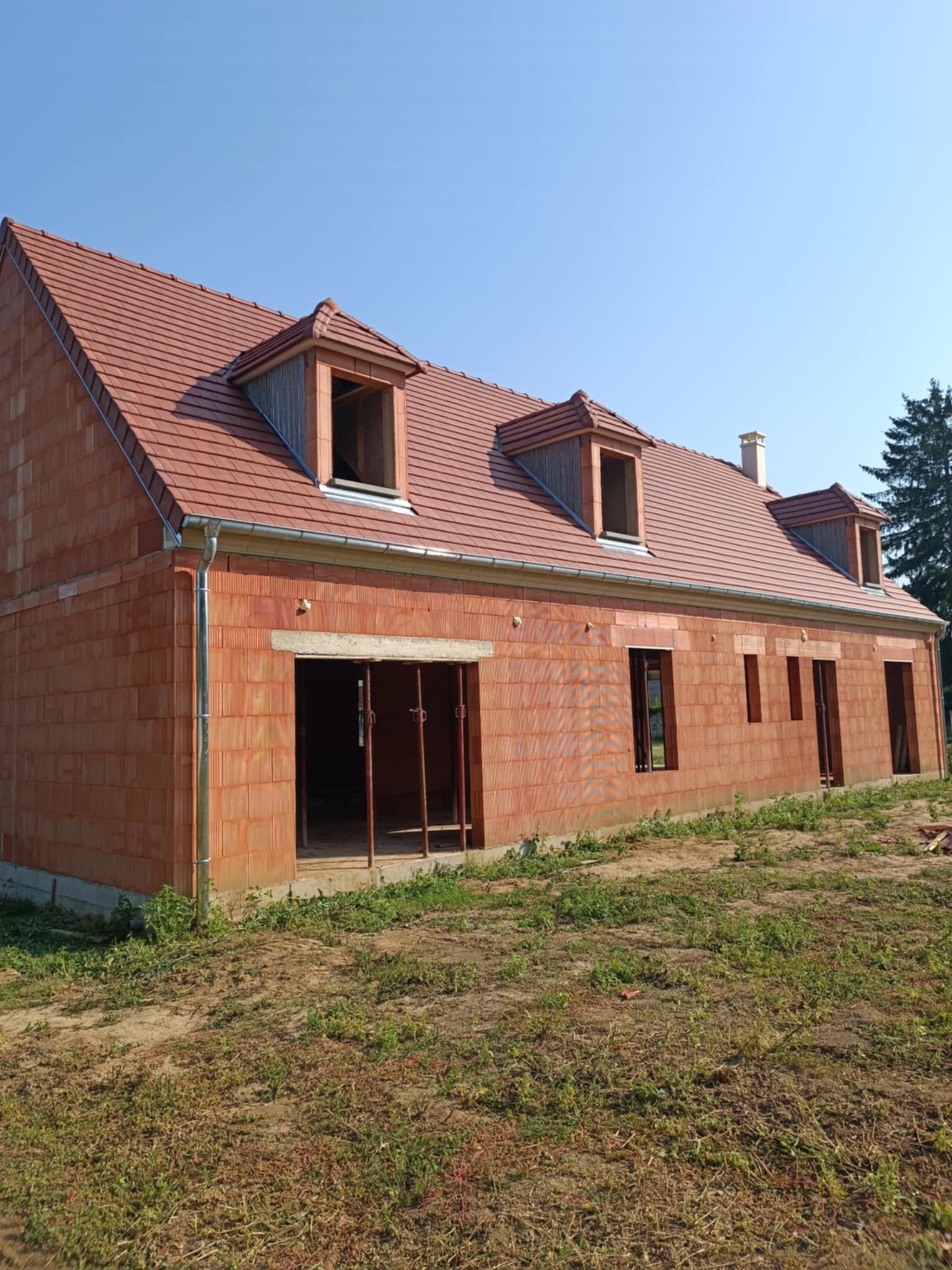 Maison en briques en construction, avec toit de tuiles rouges et lucarnes, par une journée ensoleillée.