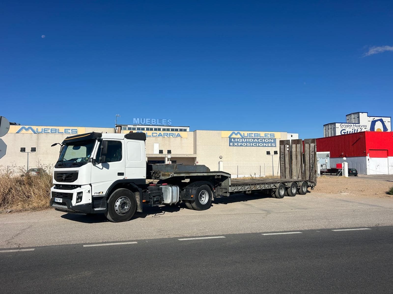 Camión semirremolque blanco con plataforma estacionado frente a un edificio con la palabra