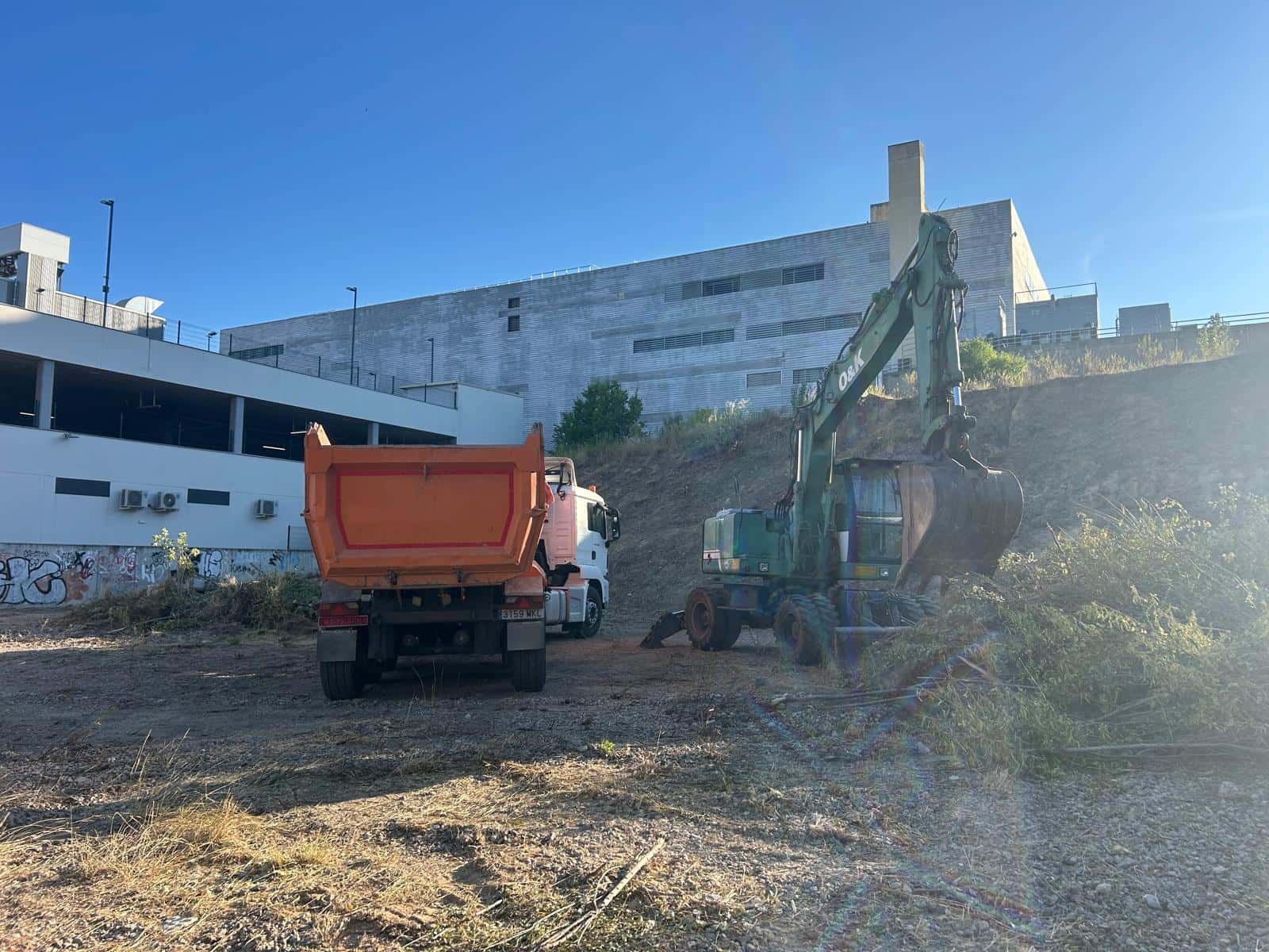 Una excavadora carga un camión volquete naranja con escombros frente a un edificio en un día soleado.